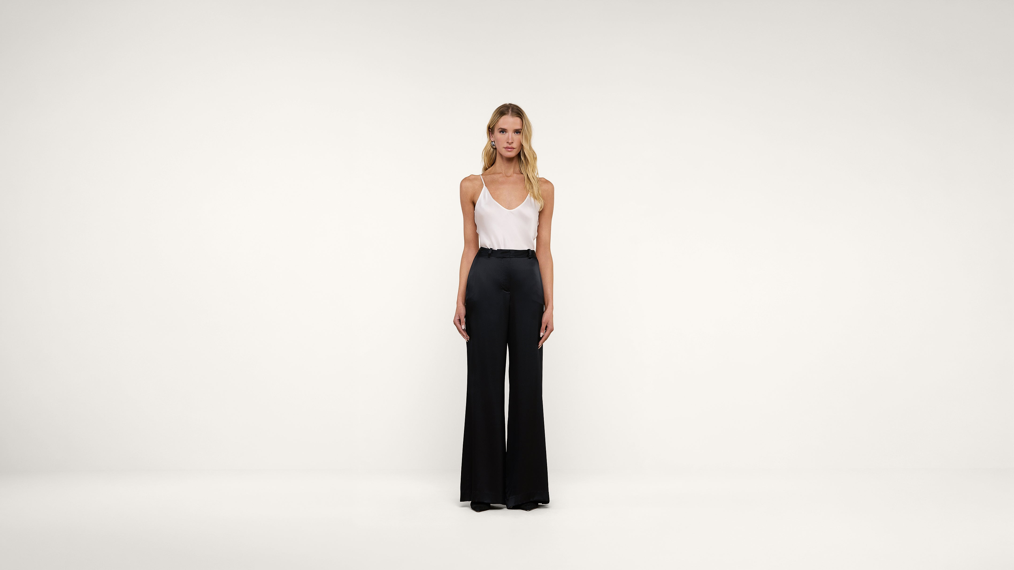 A woman stands against a plain white background wearing a white sleeveless top and high-waisted black wide-leg pants. She faces forward with her arms relaxed at her sides.