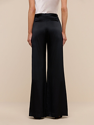 A person is shown from the back, wearing a white top and high-waisted black Pilar Silk Wide-Leg Pant with belt loops and welt pockets. The background is plain beige.