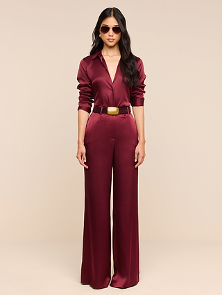 A woman wearing the Pilar Silk Wide-Leg Pant in shiny burgundy, paired with a matching high rise top with long sleeves, a wide belt, and sunglasses, stands against a plain beige background.
