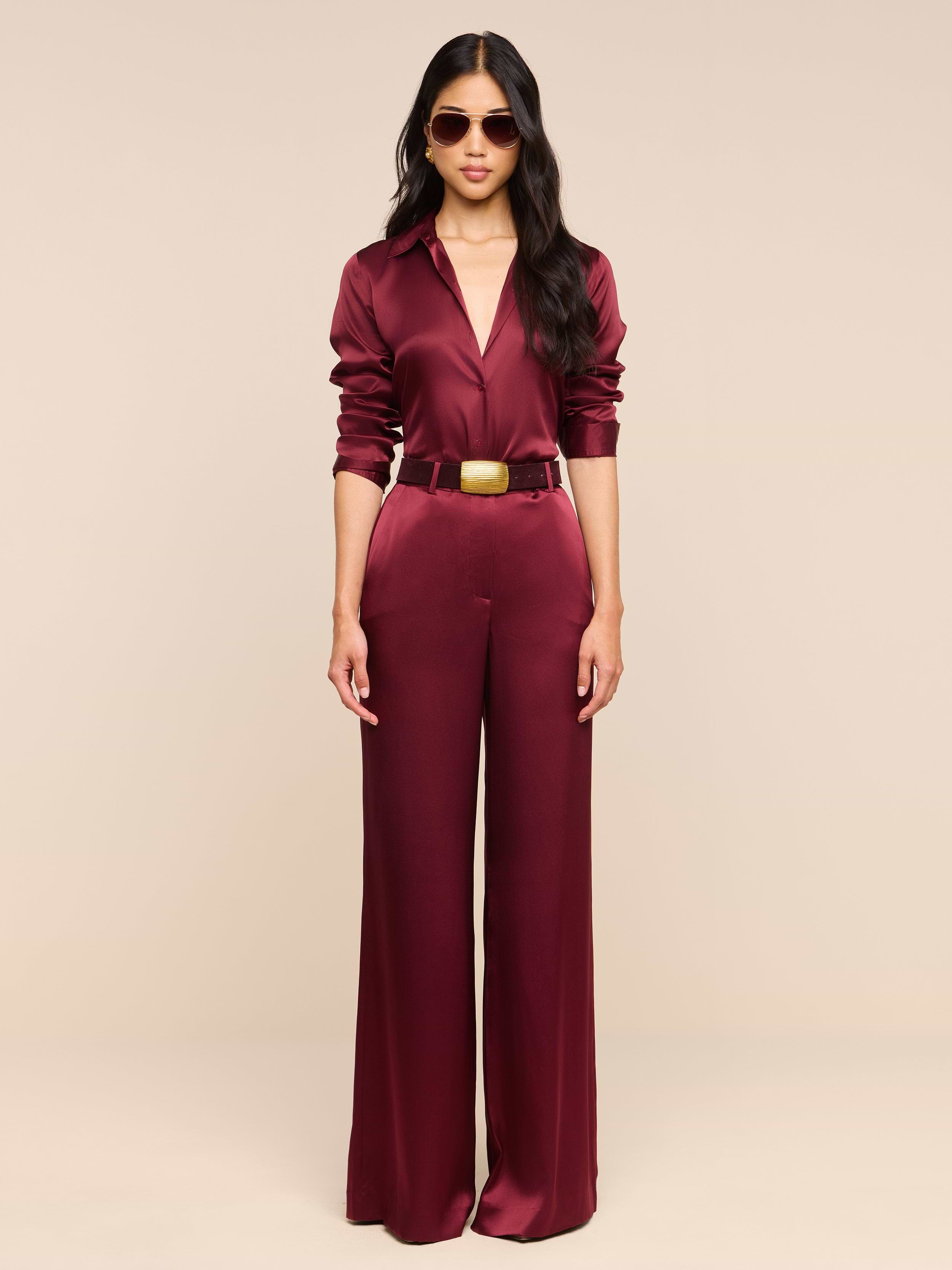 A person wearing the Pilar Silk Wide-Leg Pant in burgundy, paired with a gold belt and sunglasses, stands against a plain white background facing forward with arms relaxed by their sides.