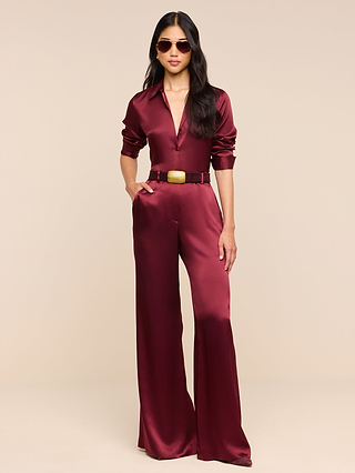 A woman stands with her hands in the pockets of the Pilar Silk Wide-Leg Pant, styled with sunglasses and a wide belt, against a plain beige background.