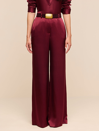 Wearing the Pilar Silk Wide-Leg Pant in maroon, styled with a matching maroon belt featuring a gold buckle, this individual stands against a plain beige background. Their ensemble includes maroon shirt sleeves that are subtly ruched at the wrists.