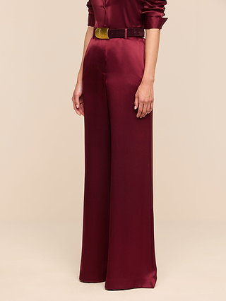 A person wearing the Pilar Silk Wide-Leg Pant in burgundy, styled with a matching belt, stands against a plain beige background. Only the lower half of the body is visible, highlighting the elegant silk wide-leg silhouette.