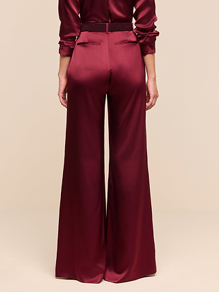 A person stands facing away, wearing the Pilar Silk Wide-Leg Pant in burgundy satin, paired with a matching long-sleeve top. The background is plain beige.