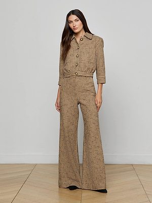 A woman with long dark hair and black shoes stands indoors against a plain wall and wooden floor, wearing Pilar Tweed Wide-Leg Pant in brown Italian windowpane tweed with a high rise, wide legs, large buttons, and a collar.