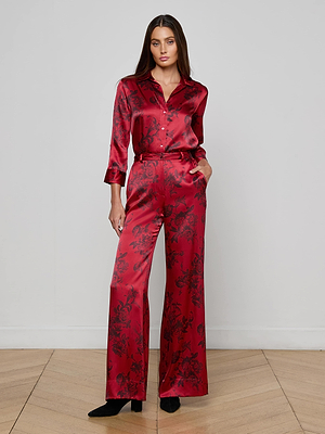 A woman with long brown hair stands against a light gray wall, wearing L'AGENCE's Pilar Silk Wide-Leg Pant in burgundy with a subtle floral print, paired with a matching silk shirt and black boots, her hands in her pockets.