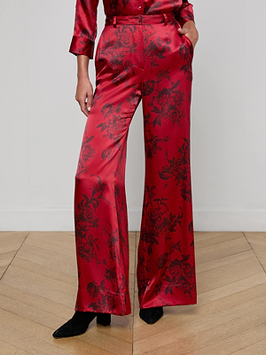 Wearing L'AGENCE Pilar Silk Wide-Leg Pant in shiny, high-waisted burgundy silk with black floral print, paired with black heeled boots and a matching red satin top, a person stands on a wood floor against a plain white wall.