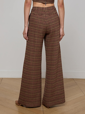 A person stands indoors on a wooden floor, wearing the L'AGENCE Pilar Houndstooth Wide-Leg Pant in brown, red, and green check with a matching cropped top. The photo is taken from behind to showcase the full length of the pants.