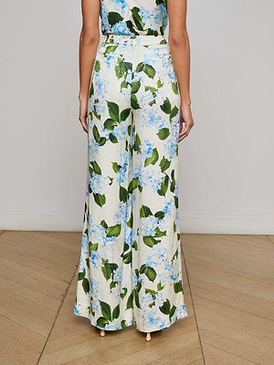 A person stands on a wooden floor, viewed from the back, wearing L'AGENCE Sale's Pilar Satin Wide-Leg Pant featuring a blue and green floral jacquard print.