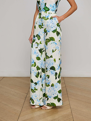 Someone wearing the L'AGENCE Sale Pilar Satin Wide-Leg Pant, featuring a blue and white hydrangea floral print, stands with hands in pockets on a wooden floor against a plain wall.