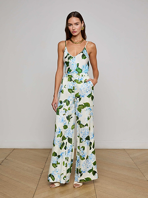 A woman models the Pilar Satin Wide-Leg Pant by L'AGENCE Sale, featuring a white base with blue and green hydrangea print. She pairs it with gold earrings and a chunky necklace, posing against a plain indoor background.