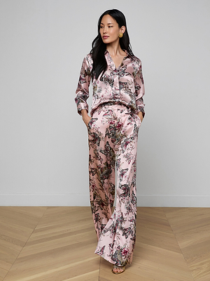 A woman stands indoors on a wooden floor wearing the Pilar Silk Wide-Leg Pant in pink as part of a pajama-style butterfly jungle print set with long sleeves, wide-leg pants, gold earrings, and her hands in her pockets. She has long black hair.