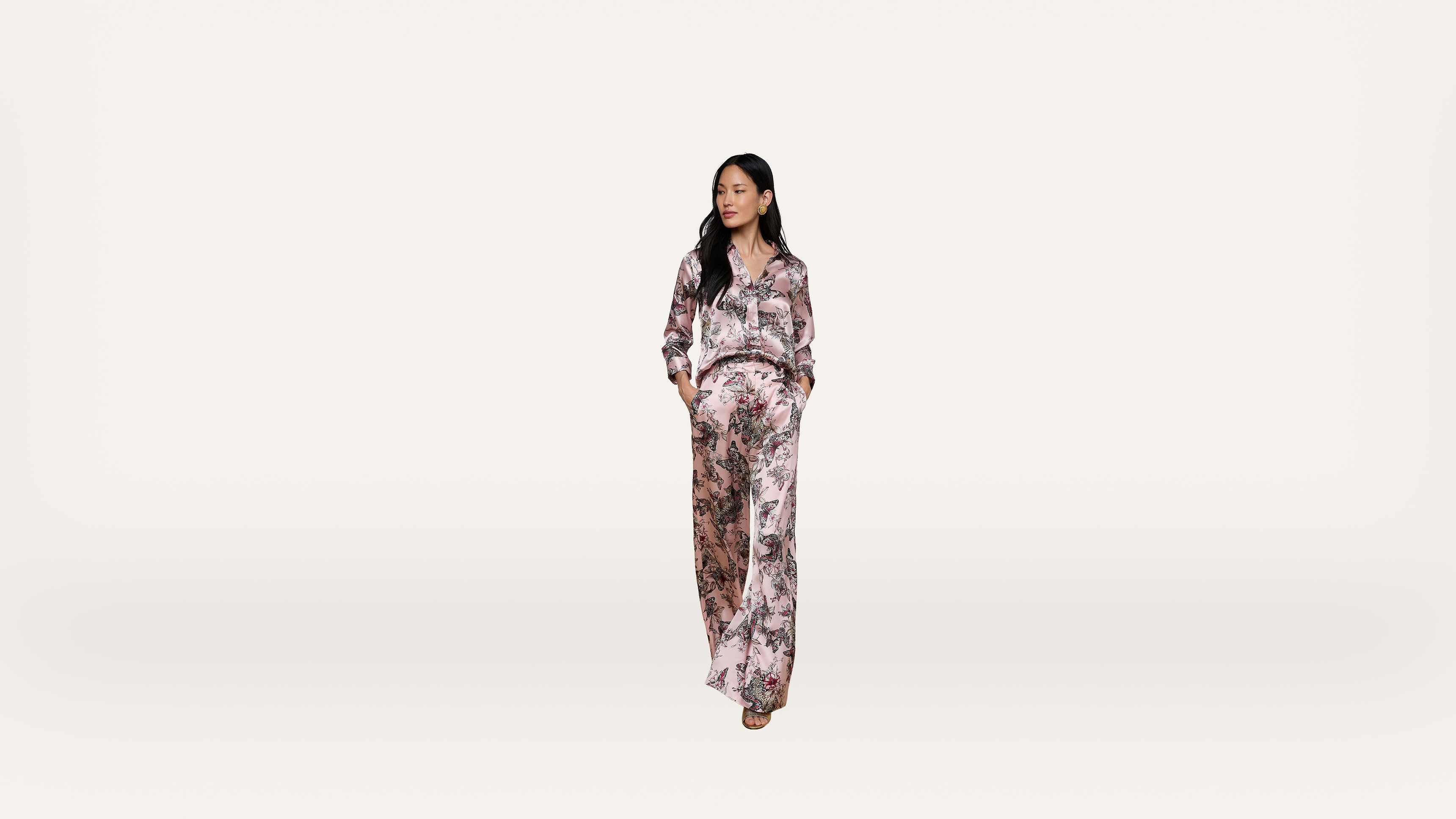 Woman standing against a plain background, wearing a loose, light-colored floral outfit with long sleeves and wide-legged pants, hands in pockets, and looking to her left.
