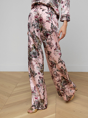A person stands on a wooden floor wearing the Pilar Silk Wide-Leg Pant in a high-rise, pink butterfly jungle print with a partially visible matching top and open-toed tan sandals.