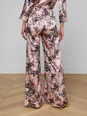 A person stands on a wooden floor wearing the Pilar Silk Wide-Leg Pant in a pink butterfly jungle print, paired with a matching long-sleeve top. The image, shot from behind, highlights the vibrant back view of the outfit.