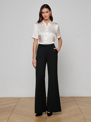 A woman stands indoors against a light wall, wearing a short-sleeve ivory satin blouse and the Pilar Wide-Leg Pant made from four-way stretch suiting. With her hands in her pockets, she faces forward in black heels.