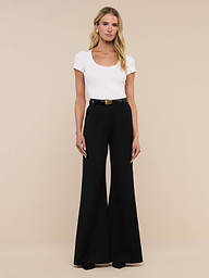 A woman stands against a plain white background, wearing a white short-sleeve top tucked into the Pilar Wide-Leg Pant, crafted from four-way stretch suiting and finished with a black belt and black shoes.