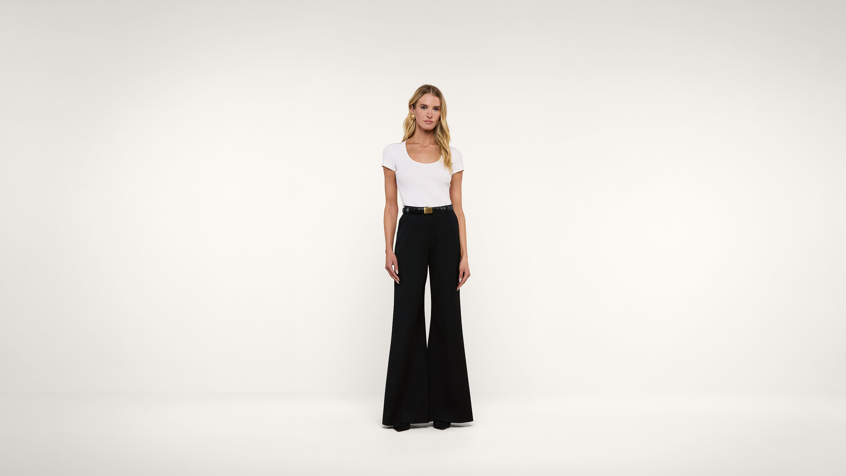 A woman stands in a white room wearing a white t-shirt tucked into high-waisted black wide-leg pants with a black belt.