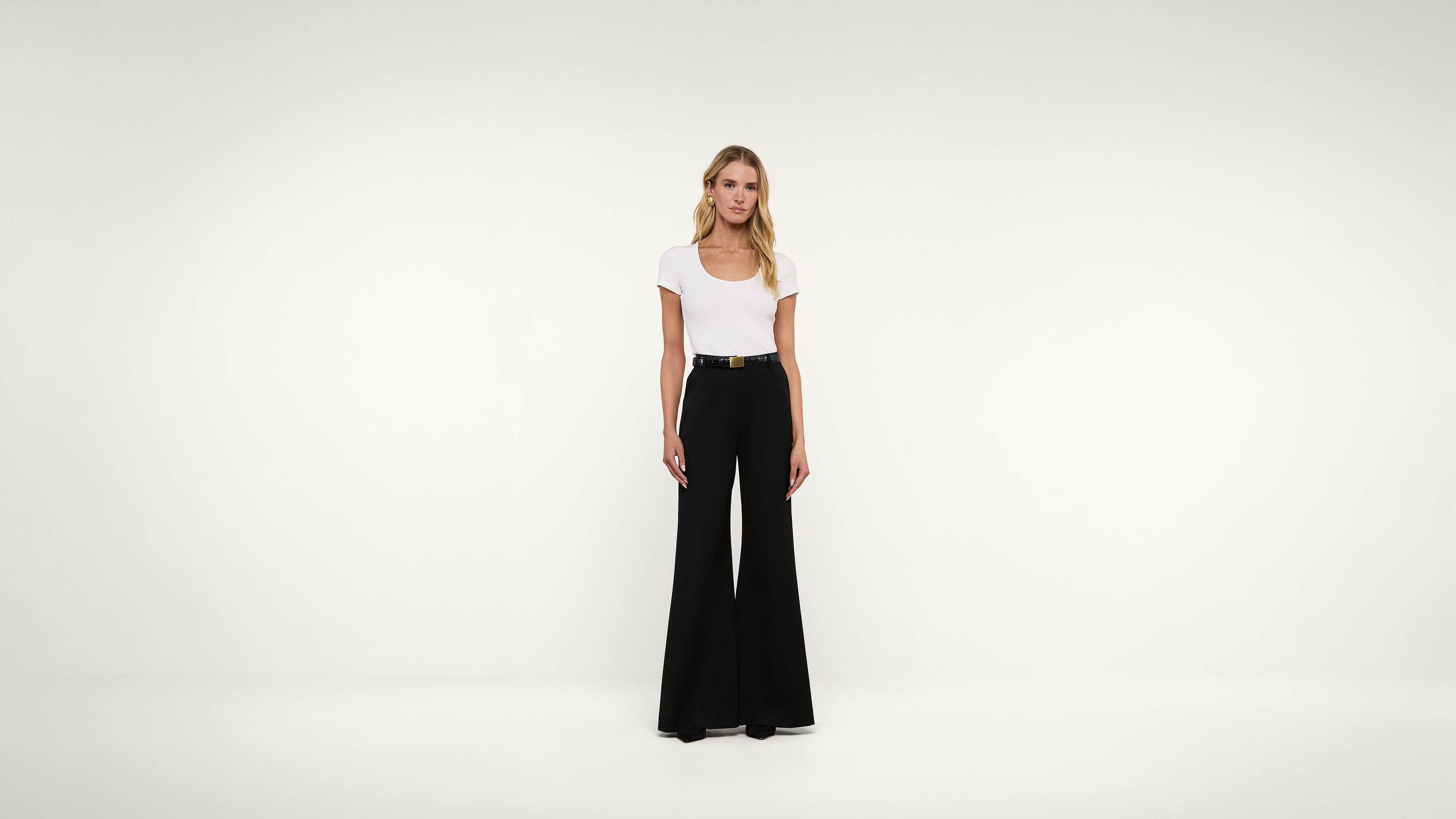 A woman stands in a white room wearing a white t-shirt tucked into high-waisted black wide-leg pants with a black belt.
