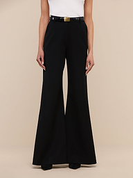 A person wearing the Pilar Wide-Leg Pant, a high-rise black flared trouser made from four-way stretch suiting, along with a white sleeveless top and black shoes stands against a plain white background. The person’s head is not visible in the image.