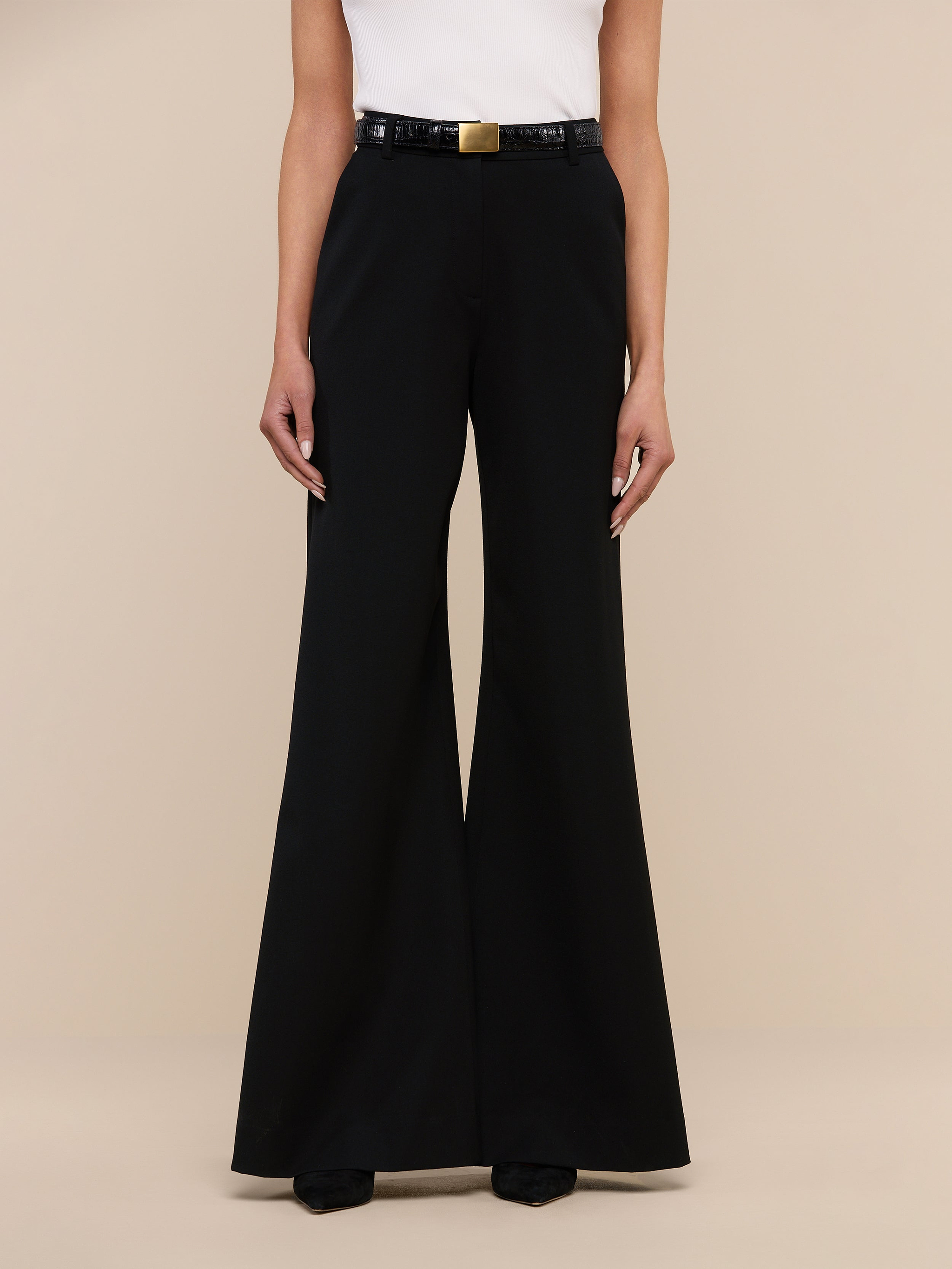A person wearing the Pilar Wide-Leg Pant, a high-rise black flared trouser made from four-way stretch suiting, along with a white sleeveless top and black shoes stands against a plain white background. The person’s head is not visible in the image.