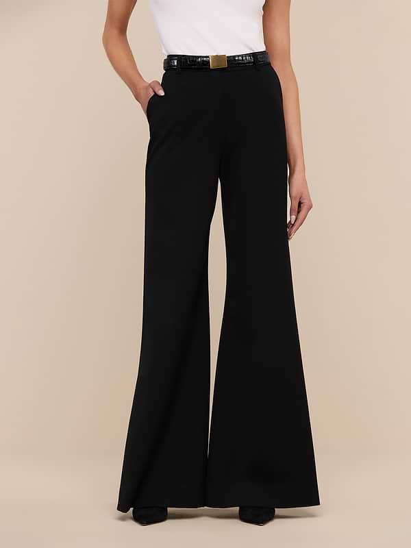 A person wearing the Pilar Wide-Leg Pant in black, crafted from four-way stretch suiting, paired with a white sleeveless top and a black belt with a gold buckle, stands against a plain white background.