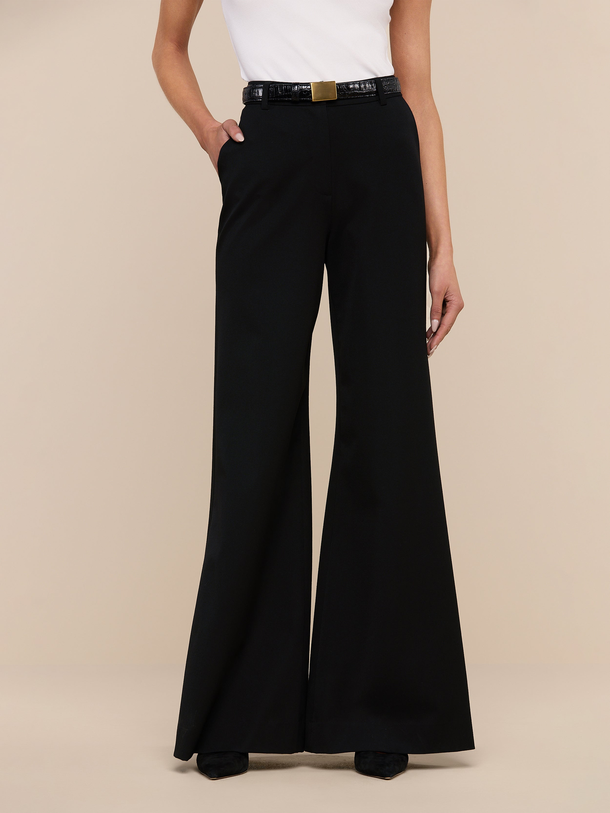 A person wearing the Pilar Wide-Leg Pant in black, crafted from four-way stretch suiting, paired with a white sleeveless top and a black belt with a gold buckle, stands against a plain white background.