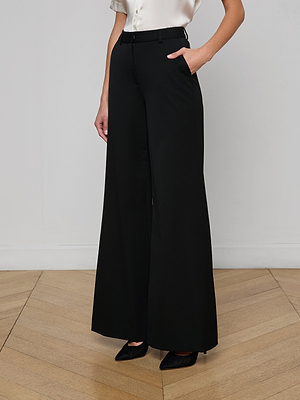 Wearing the Pilar Wide-Leg Pant in suiting fabric, paired with a white blouse and black pointed-toe heels, a person stands on a light wooden floor with one hand in their pocket.