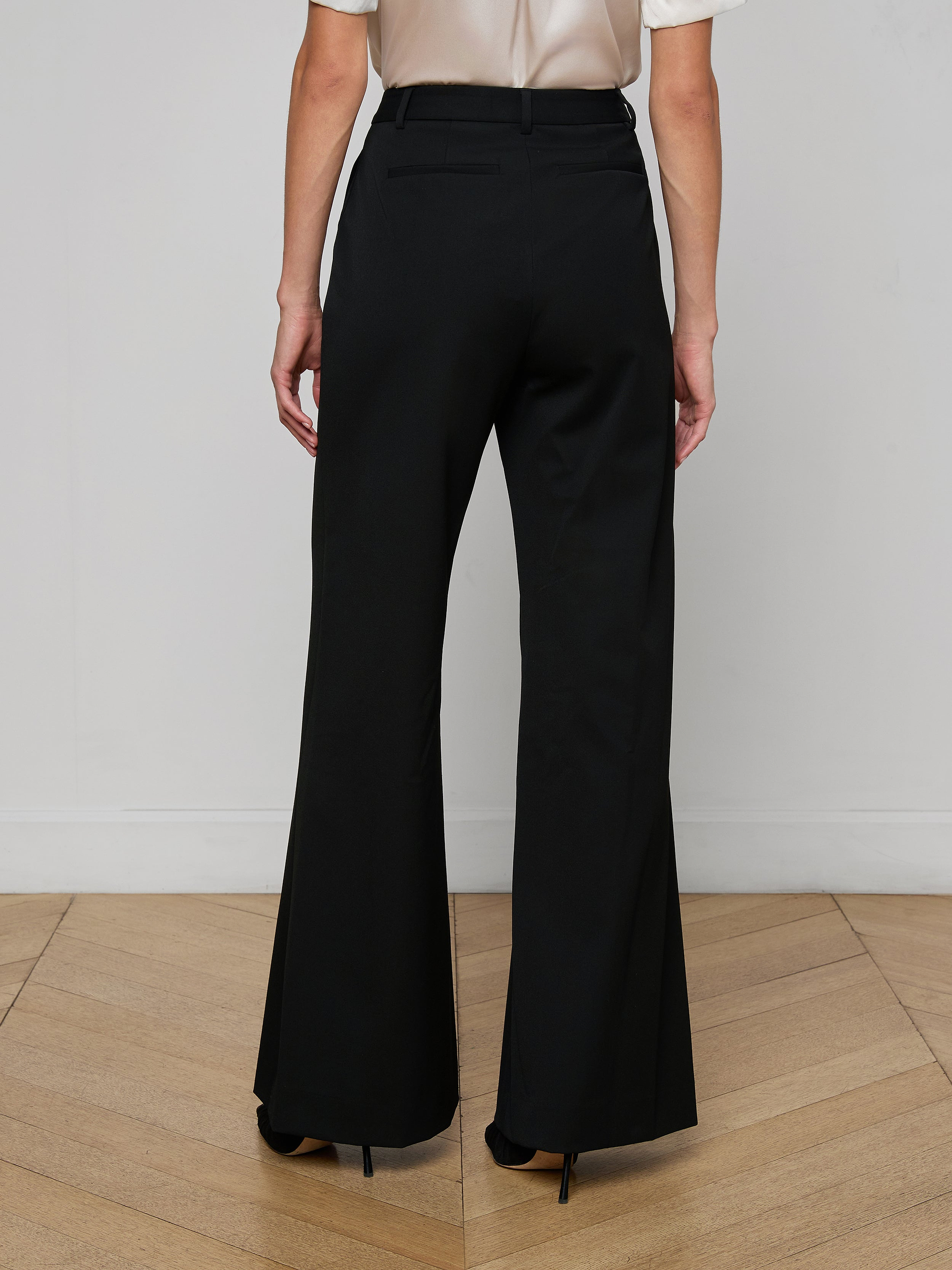 A person stands with their back to the camera, wearing the Pilar Wide-Leg Pant in black and a white top, set against a plain white background.