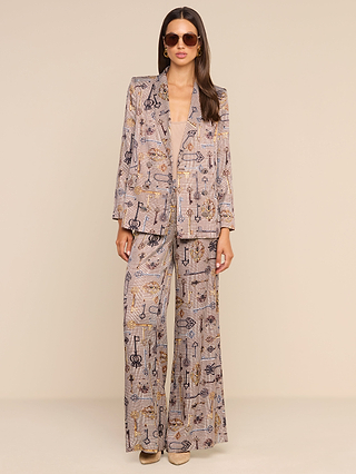 A woman wearing sunglasses, a patterned blazer, and the Pilar Wide-Leg Pant in brown houndstooth with beige shoes stands against a plain light background.