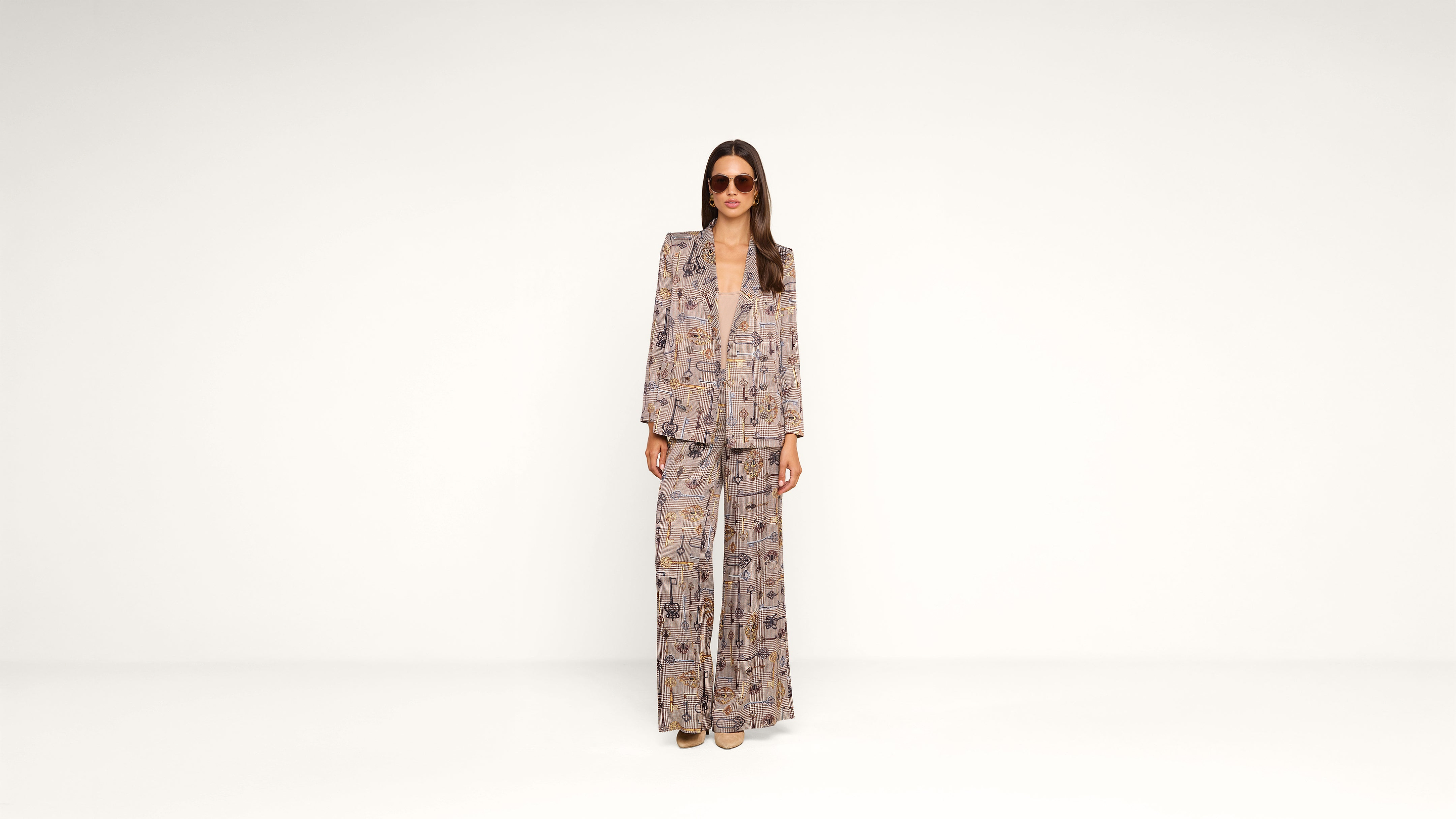 A woman stands against a plain white background wearing a patterned suit with a blazer and wide-leg pants, sunglasses, and brown shoes.