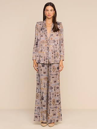 A woman stands against a neutral background wearing the Pilar Wide-Leg Pant, paired with a matching patterned, loose-fitting blazer featuring abstract designs. She has long brown hair, gold earrings, and beige heels.
