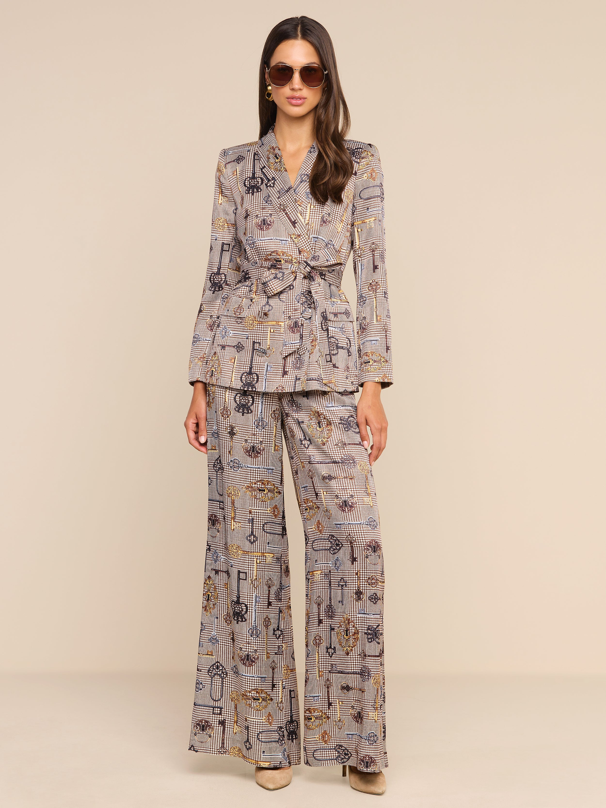 A woman stands against a plain white background wearing sunglasses and a matching printed suit, featuring the Pilar Wide-Leg Pant in a brown houndstooth pattern and a belted blazer.
