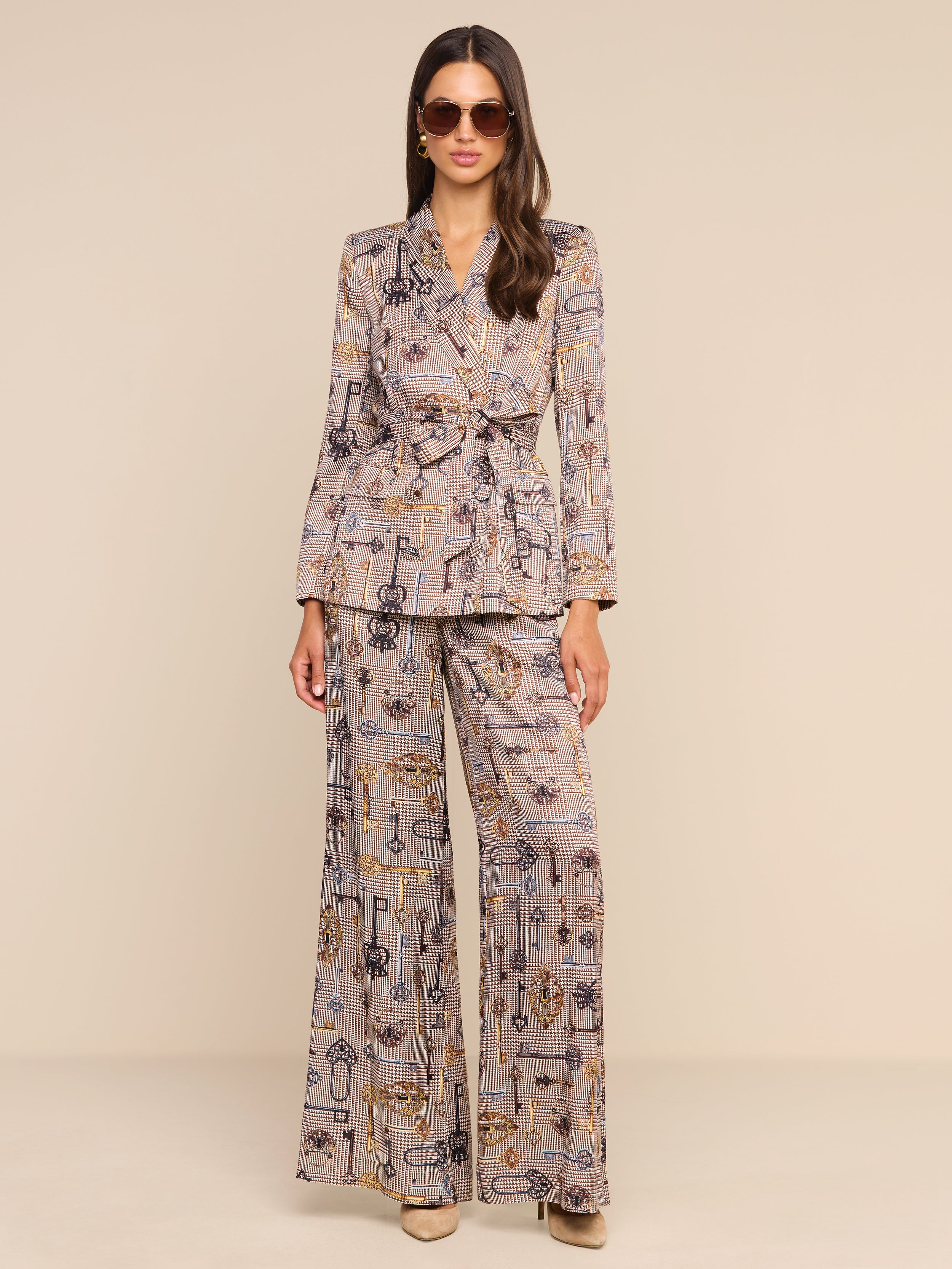 A woman stands against a plain white background wearing sunglasses and a matching printed suit, featuring the Pilar Wide-Leg Pant in a brown houndstooth pattern and a belted blazer.