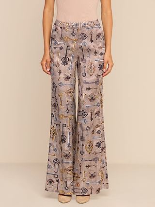 A woman wears the Pilar Wide-Leg Pant, featuring a high-waisted, wide-leg silhouette and a luxurious satin jacquard fabric adorned with a beige, black, and gold geometric and key print. She pairs it with a light pink top and beige shoes while standing against a plain background.