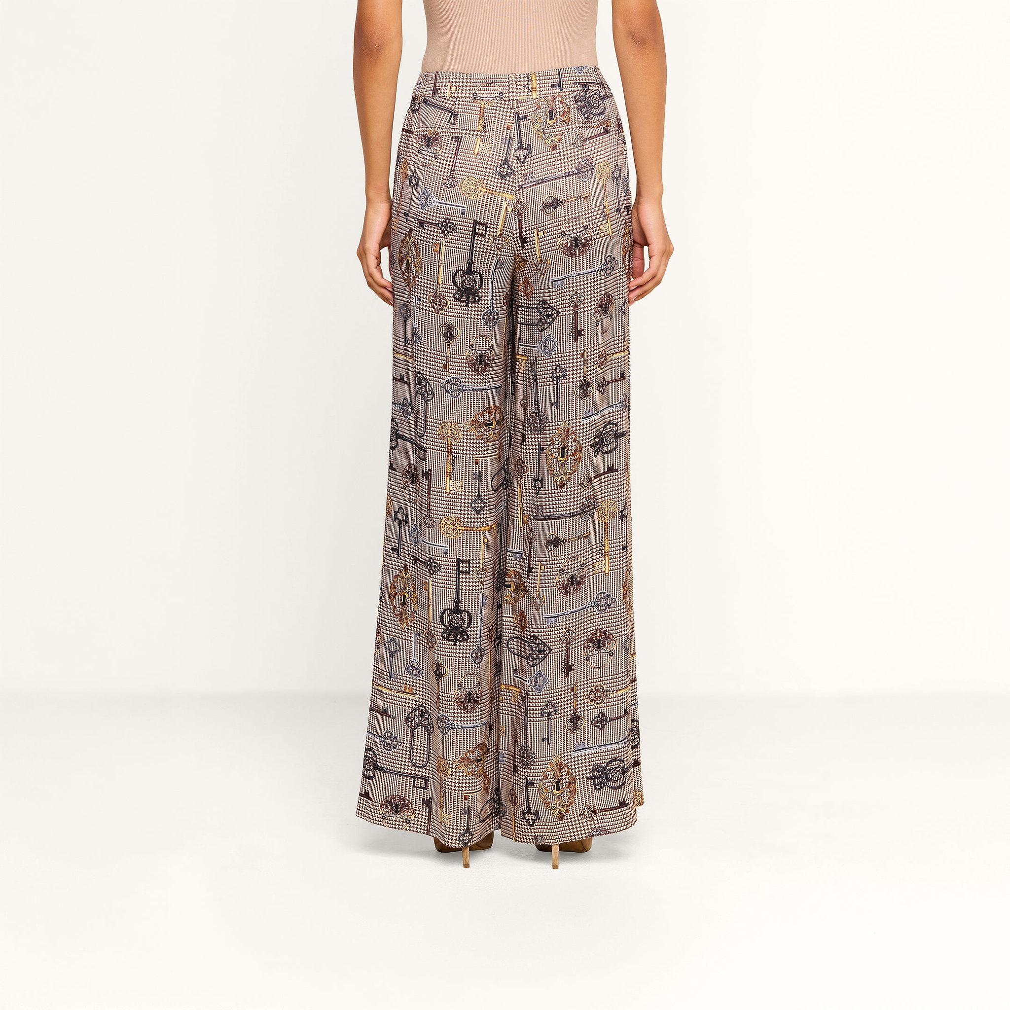 A person stands facing away, wearing the Pilar Wide-Leg Pant featuring various abstract designs, paired with a fitted, sleeveless beige top against a plain white background.