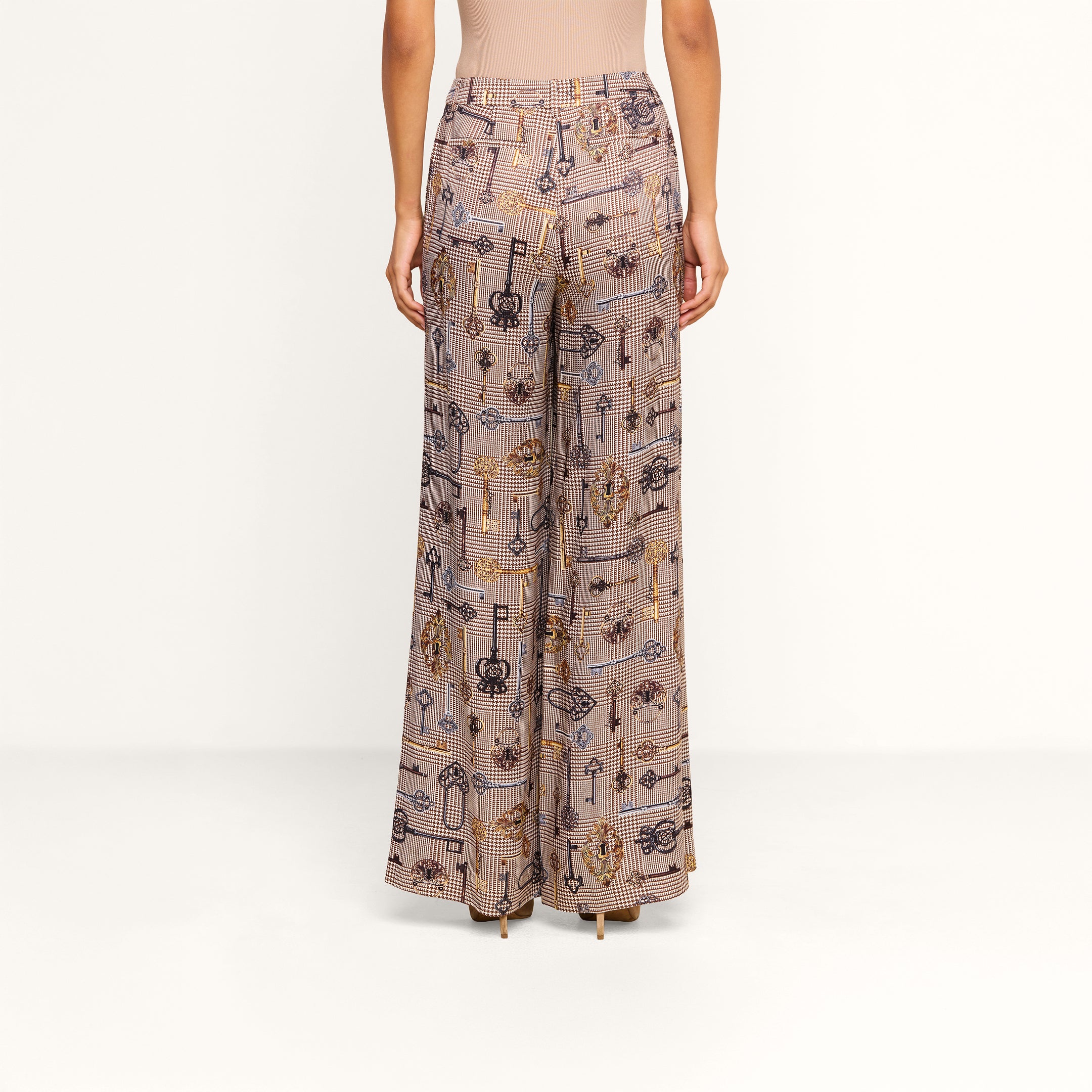 A person stands facing away, wearing the Pilar Wide-Leg Pant featuring various abstract designs, paired with a fitted, sleeveless beige top against a plain white background.