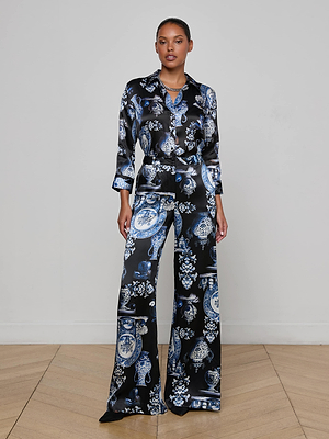 A woman stands against a plain wall wearing the L'AGENCE Pilar Silk Wide-Leg Pant, featuring a blue and white porcelain print on black. She faces forward with a neutral expression, paired with a matching satin shirt.