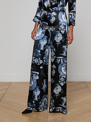 A person wears the Pilar Silk Wide-Leg Pant by L'AGENCE, featuring a blue and white vase-inspired print, paired with a matching long-sleeve top. The shiny set is styled on a polished wooden floor.