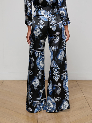 A person stands on a wooden floor wearing L'AGENCE Pilar Silk Wide-Leg Pants with a blue and white porcelain print, shown from the waist down facing a plain white wall.