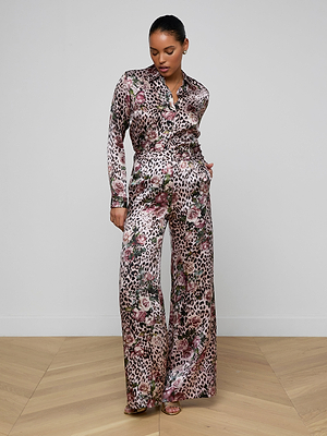 A woman stands on a wooden floor in Pilar Silk Wide-Leg Pant, featuring a rose cheetah and floral print. She looks downward with her hands in her pockets, set against a plain white wall.