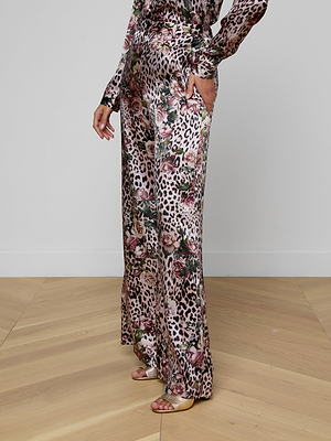 A person stands on a wooden floor wearing the Pilar Silk Wide-Leg Pant in a rose cheetah print, paired with a matching long-sleeve top. Only the lower half of their body is visible.