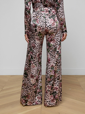 Someone stands on a light wood floor, seen from behind, wearing the Pilar Silk Wide-Leg Pant featuring a rose cheetah print in pink, green, and brown. The photo highlights their full legs and part of their torso.