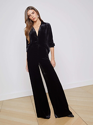 A woman wearing high-rise black Pilar Velvet Wide-Leg Pants and a matching long-sleeve top stands against a plain light background, looking slightly to the side.