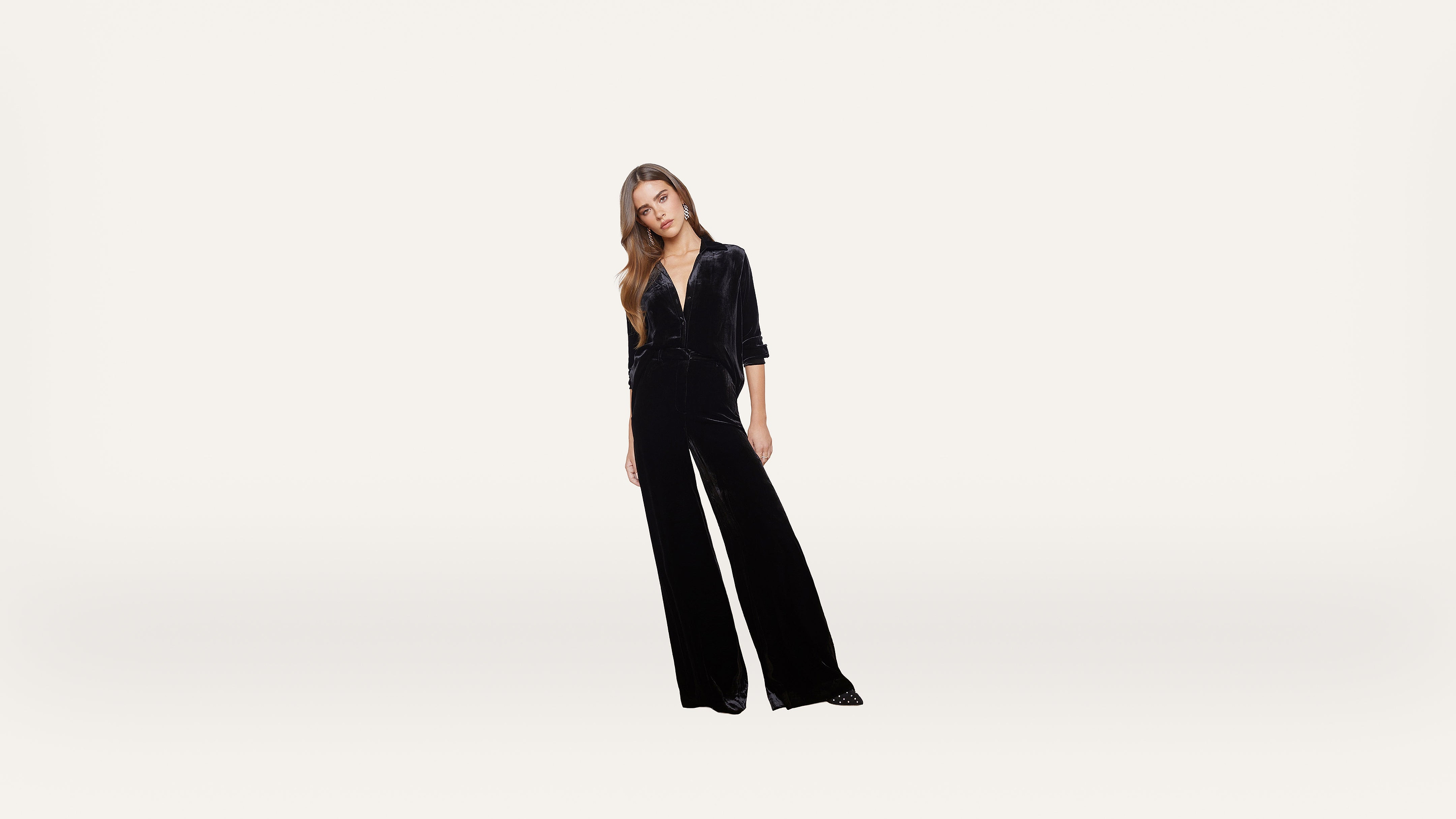 A woman wearing a black velvet jumpsuit stands against a plain white background, with her head slightly tilted and hands relaxed by her sides.