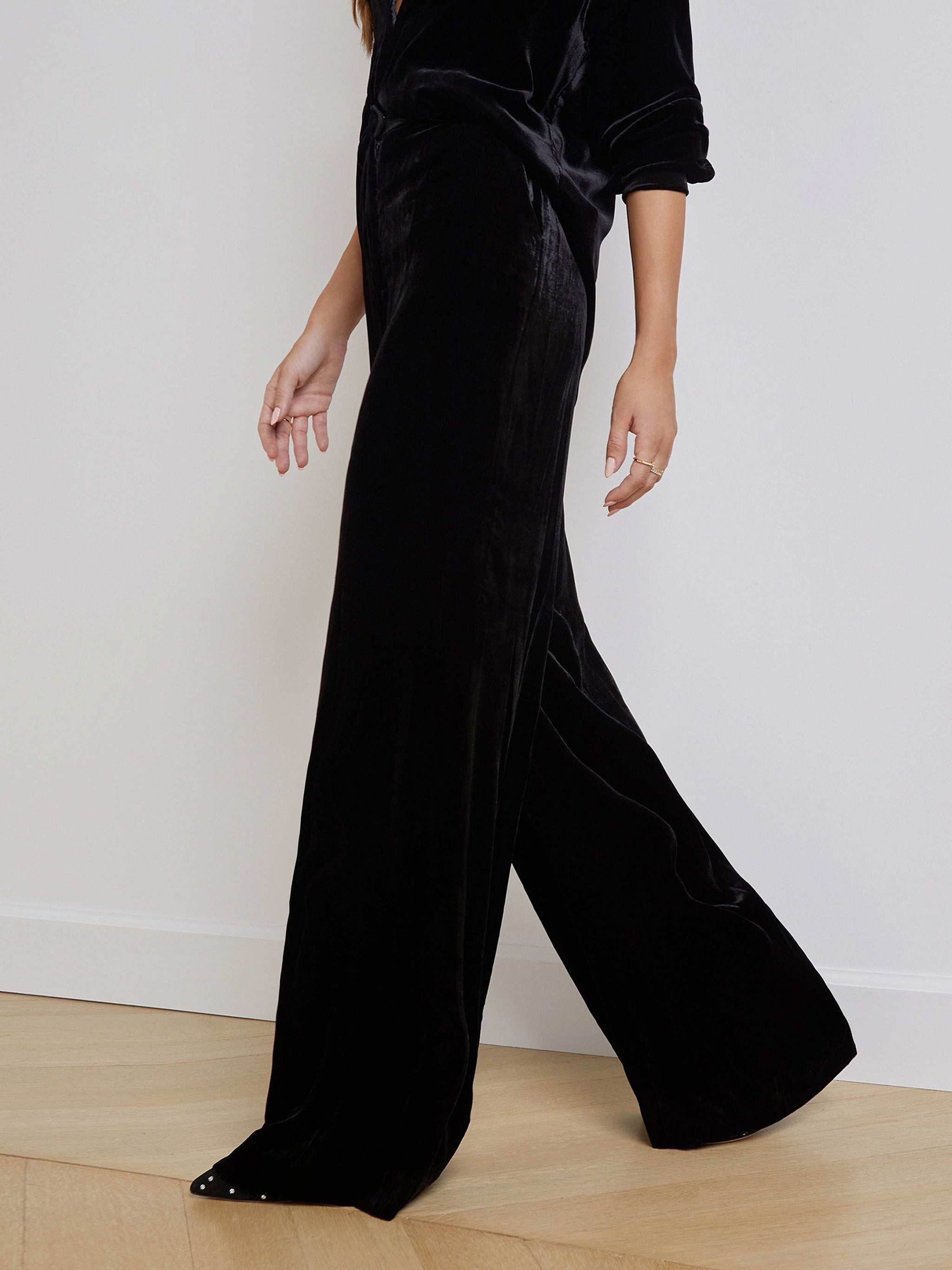 A person shown from the shoulders down walks against a plain white background, wearing the black Pilar Velvet Wide-Leg Pant with a matching top.