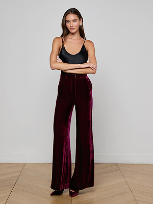 A woman with long brown hair stands with arms crossed, wearing the L'AGENCE Pilar Velvet Wide-Leg Pant in burgundy and a black spaghetti strap top, posed against a plain light gray wall on a wooden floor.