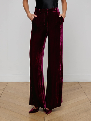 Dressed in the L'AGENCE Pilar Velvet Wide-Leg Pant, a person stands on a wooden floor against a white wall with hands in their pockets, showcasing the high-rise, burgundy velvet pants paired with pointed-toe shoes.
