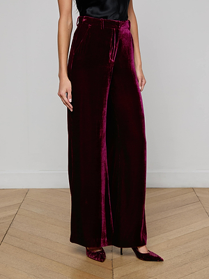 A person stands on a light wood floor against a white wall, wearing the L'AGENCE Pilar Velvet Wide-Leg Pant in deep burgundy, paired with matching pointed-toe heels and a black sleeveless top.