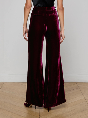 Someone wearing L'AGENCE Pilar Velvet Wide-Leg Pants in burgundy and black high heels stands on a light wooden floor against a plain white wall.